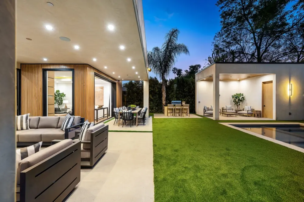Modern outdoor patio area with comfortable seating, dining table, and a lush green lawn, set against a twilight sky.