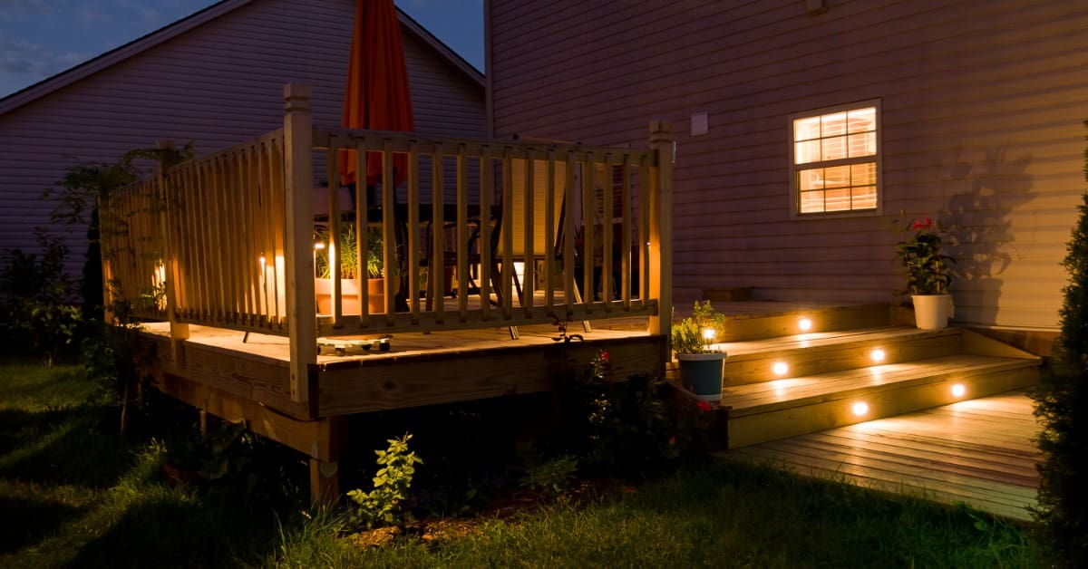 A wooden deck against the backside of a house has a table, chairs, closed umbrella, and step lighting.
