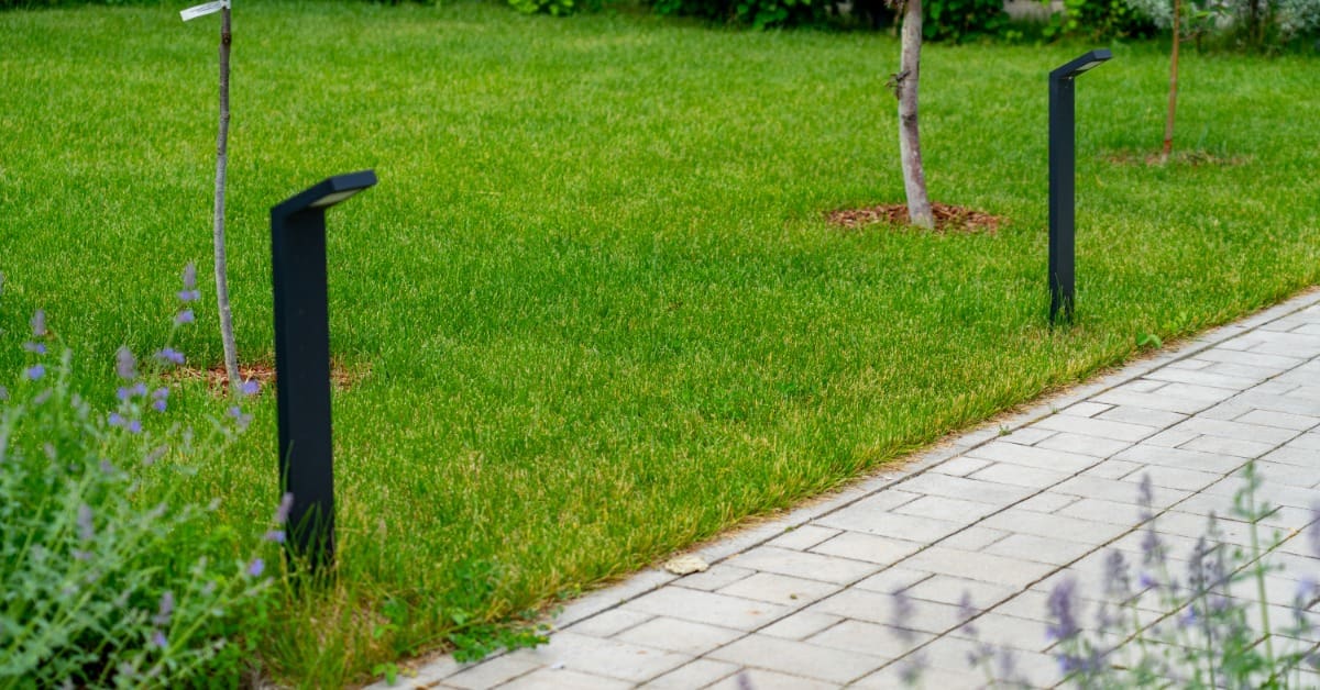 Two sleek black bollard lights installed along a stone paver walkway. Small trees and bushes are in the background.