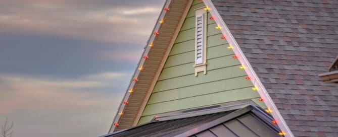 The upper level of a house with green siding has permanent yellow and red holiday lights installed on the roofline.