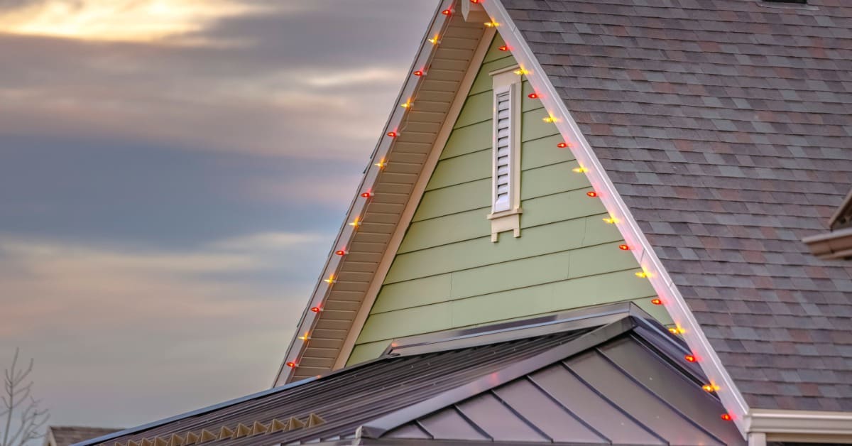 The upper level of a house with green siding has permanent yellow and red holiday lights installed on the roofline.