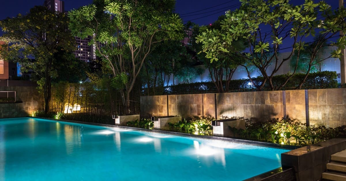 A backyard with a clear blue swimming pool and tall trees. Lights are positioned along the pool and the stone fence.