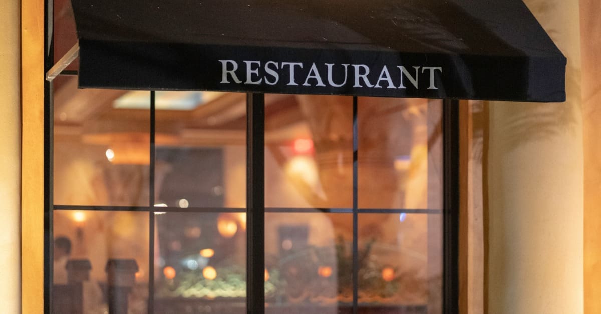 A black canopy awning with the text "Restaurant" is above a window. Warm lights brighten the exterior and interior.