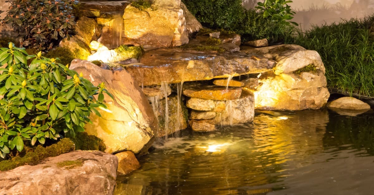 A small backyard waterfall with layers of large and small rocks. Staggered spotlights illuminate the waterfall and pond.