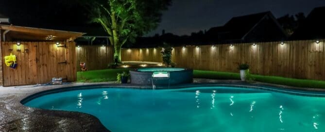 A curved pool and hot tub in a backyard is surrounded by concrete landscaping. Lights line the top of the wooden fence.