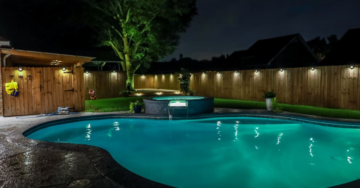 A curved pool and hot tub in a backyard is surrounded by concrete landscaping. Lights line the top of the wooden fence.