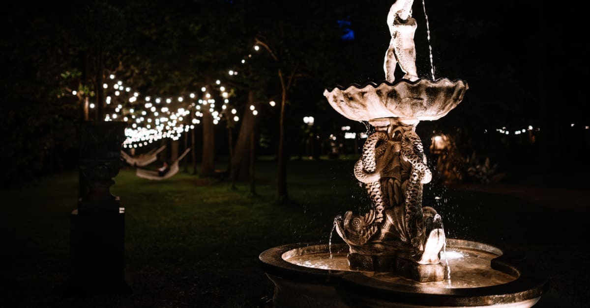 A tall water fountain has accent lights at the top and base. String lights illuminate a seating area in the background.