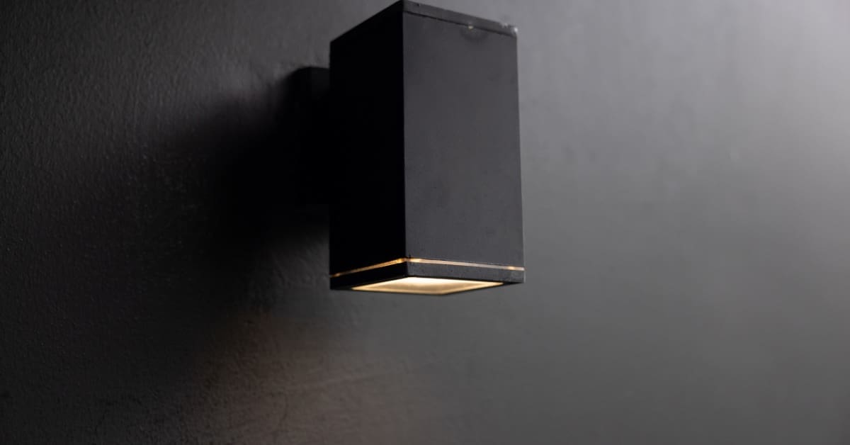 A black, rectangular light fixture is mounted on a smooth black wall. White light glows from the base of the fixture.
