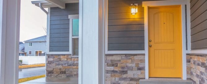 The entryway of a home with a yellow door, a glowing wall-mounted lantern, and a damp concrete porch.