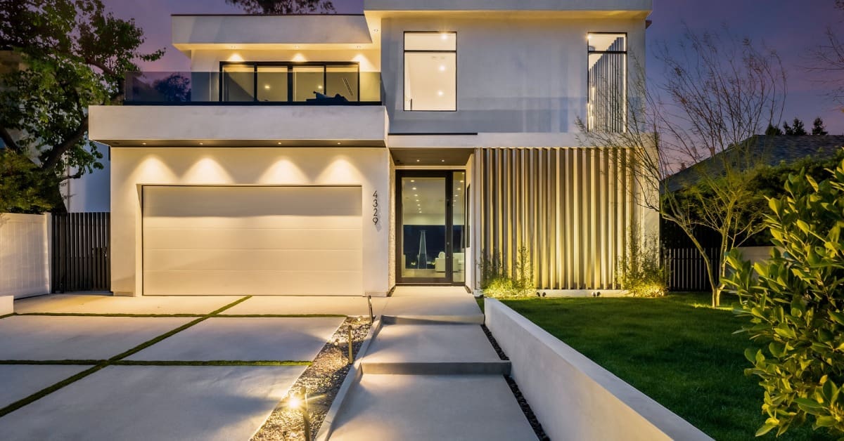 A modern house exterior is glowing at night with a concrete driveway, lawn, warm entry lighting, and lit windows.