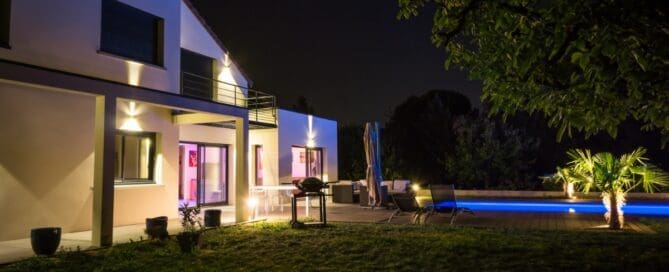 A large modern house glowing at night with poolside seating, palm trees, and exterior accent lighting.