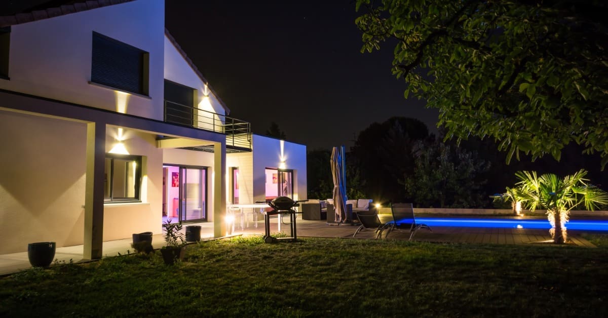 A large modern house glowing at night with poolside seating, palm trees, and exterior accent lighting.
