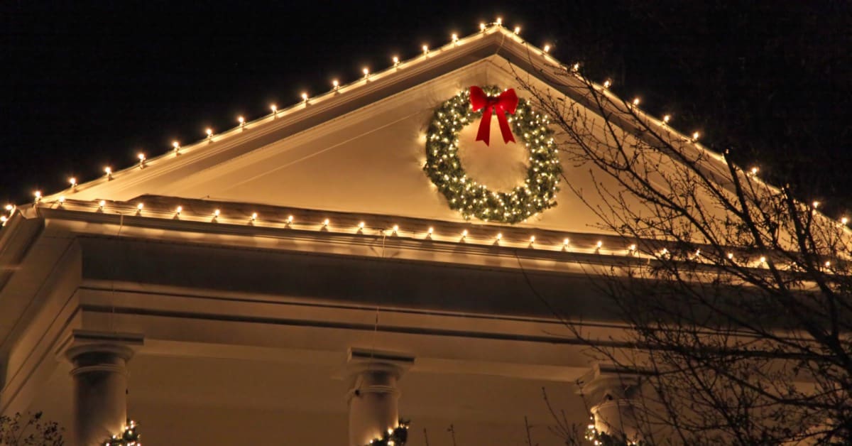 A home at night with an illuminated wreath and white lights on the roof. Garland with lights is wrapped around the columns.