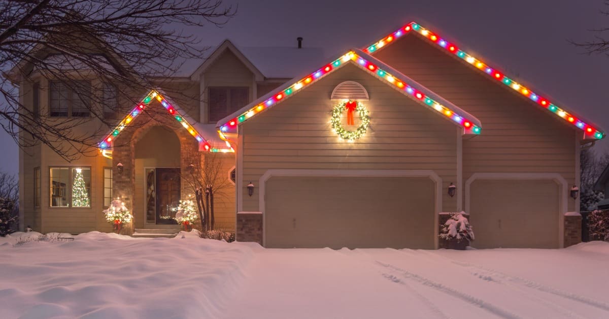 A home with a snow-covered yard and driveway has multicolored lights lining the three gables on the roof.